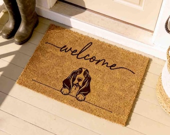 May include: A rectangular tan coir doormat featuring the word welcome in black cursive script above a black line drawing of a Basset Hound dog. The mat is positioned on a light-colored porch floor in front of a door with two dark rubber boots.