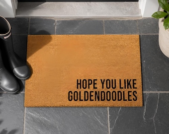 May include: A rectangular tan coir doormat with black text printed in the bottom right corner that reads, "HOPE YOU LIKE GOLDENDOODLES." The mat has a coarse, fibrous texture and is positioned on a dark gray tiled floor near a doorway.