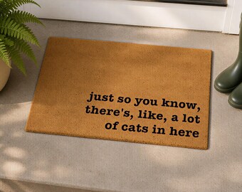 May include: A rectangular tan coir doormat with black text that reads, "just so you know, there's, like, a lot of cats in here." The mat has a coarse, fibrous texture and is positioned on a concrete step near a green plant and boots.