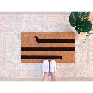 A person's feet standing on a patterned rug, with a black and brown dachshund-shaped doormat featuring a black line design.