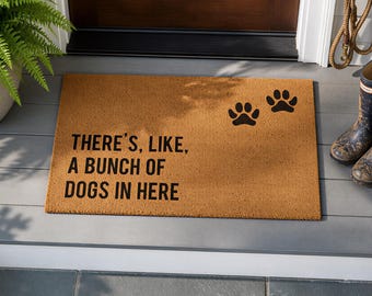 May include: A rectangular tan coir doormat featuring the black printed text THERE'S, LIKE, A BUNCH OF DOGS IN HERE and two black paw prints. The mat has a coarse, fibrous texture and is positioned on a grey porch surface in front of a dark door.