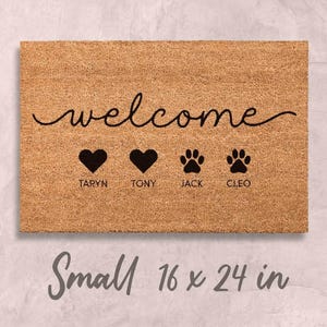 A welcome mat with the word "welcome" written on it, along with three heart symbols and the names "TARYN, TONY, JACK, CLEO". The mat is described as small, measuring 16 x 24 inches.