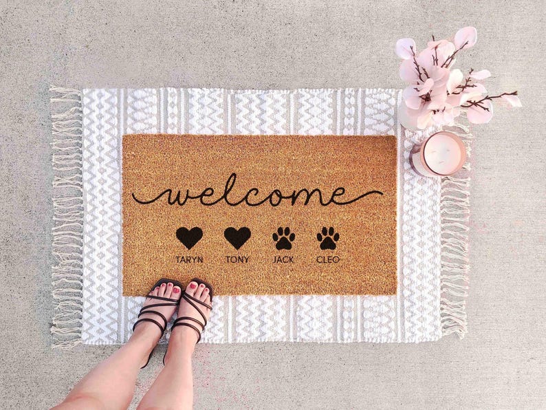 A welcome mat with the word "welcome" and three heart symbols, along with the names "TARYN", "TONY", and "JACKA" written below. The mat is placed on a white and gray patterned rug, and there are a pair of feet and a vase with pink flowers nearby.