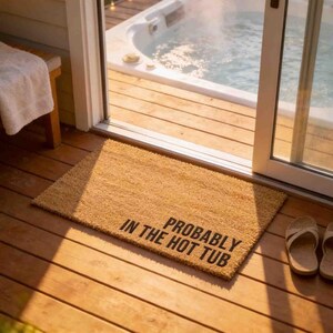 A wooden deck with a doormat that says "Probably in the hot tub" in bold letters. A hot tub is visible through a sliding glass door, and a pair of sandals is placed on the floor next to the doormat.