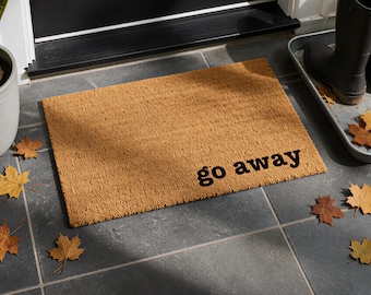 May include: A rectangular tan coir doormat with a coarse, fibrous texture. The words "go away" are printed in a bold, black, lowercase sans-serif font in the bottom right corner of the mat, creating a clear contrast against the tan surface.