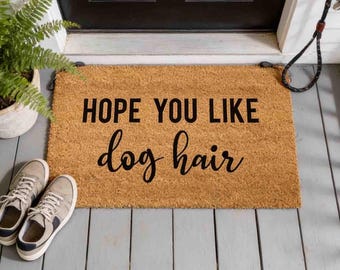 May include: A rectangular tan coir doormat featuring the black printed text HOPE YOU LIKE dog hair. The mat is positioned on a gray wooden porch floor in front of a black door, with a potted green fern and a pair of gray sneakers visible nearby.