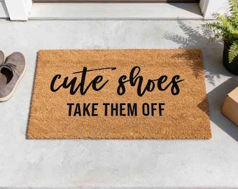 May include: A rectangular tan coir doormat featuring the black text "cute shoes" in a cursive script font above the words "TAKE THEM OFF" in a bold, sans-serif capital letter font. The mat has a coarse, natural fiber texture.