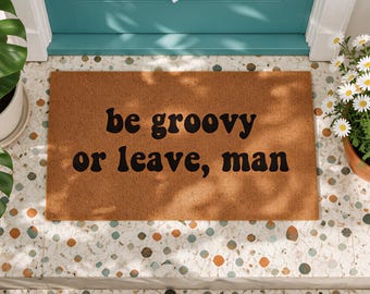 May include: A rectangular brown coir doormat featuring the black text "be groovy or leave, man" printed in a bold, rounded font. The mat has a coarse, fibrous texture and is positioned on a light-colored speckled floor surface.