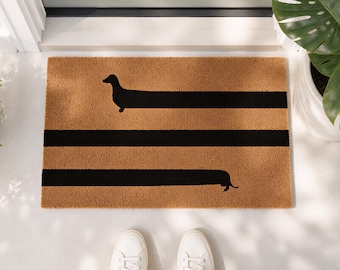 May include: A rectangular tan coir doormat featuring a black graphic of a long dachshund dog. The dog's body is elongated into two horizontal black stripes that span across the width of the mat, with the head on the left and tail on the right.