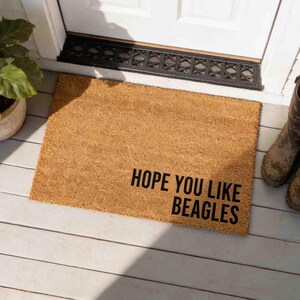 May include: A rectangular tan coir doormat with a coarse, fibrous texture. The mat features the black printed text "HOPE YOU LIKE BEAGLES" positioned in the lower right corner, written in a bold, sans-serif, all-caps typeface.