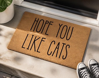 May include: A rectangular tan coir doormat with black text that reads HOPE YOU LIKE CATS in a simple sans-serif font. The mat is positioned on a concrete step in front of a dark door, with a portion of a potted plant visible in the upper left corner.