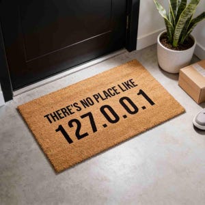 May include: A rectangular tan coir doormat featuring the black printed text THERE'S NO PLACE LIKE 127.0.0.1. The mat has a coarse, natural fiber texture and is positioned on a light gray floor in front of a dark door and a potted green plant.