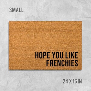 A rectangular, light brown, textured doormat with the text "HOPE YOU LIKE FRENCHIES" printed on it.