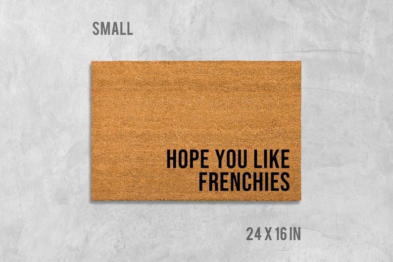 A rectangular, light brown, textured doormat with the text "HOPE YOU LIKE FRENCHIES" printed on it.