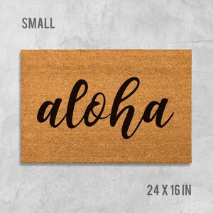 A rectangular, light brown, textured doormat with the word "aloha" written on it in a cursive font.