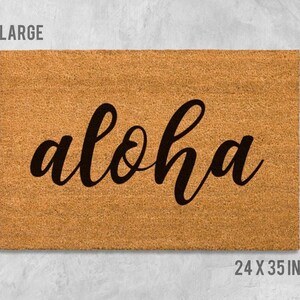 A large, rectangular, beige-colored doormat with the word "aloha" written on it in a cursive font.
