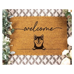 A welcome mat with a dog silhouette and the word "welcome" written on it, surrounded by greenery and flowers.