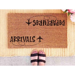 A pair of feet standing on a beige rug with a decorative mat that says "Arrivals" and has a symbol of an airplane on it.