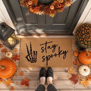 A Halloween-themed door mat with the text "Stay spooky" and a skeleton hand graphic, surrounded by fall decorations such as pumpkins, gourds, and a wreath.