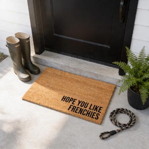 a door mat with the text "HOPE YOU LIKE FRENCHIES" on it, along with a pair of black boots and a potted plant.