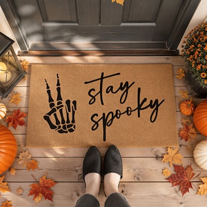 A pair of feet standing on a mat with the text "Stay Spooky" and a skeleton hand graphic, surrounded by fall decorations such as pumpkins and autumn leaves.