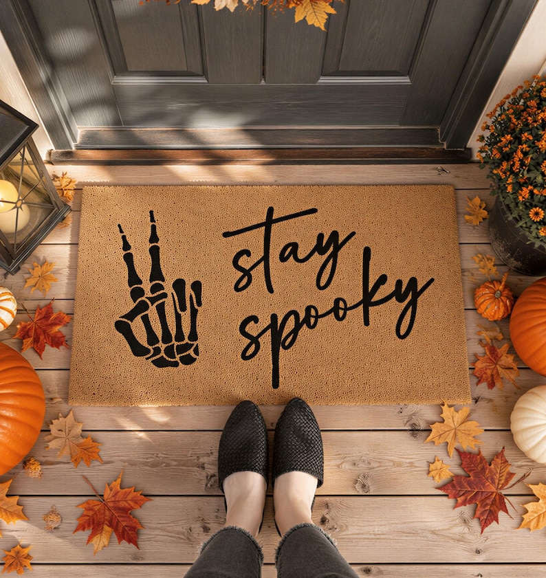 A pair of feet standing on a mat with the text "Stay Spooky" and a skeleton hand graphic, surrounded by fall decorations such as pumpkins and autumn leaves.