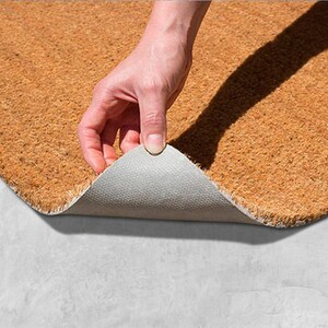 A hand is holding a piece of a cork or natural fiber mat, which is being stretched out on a concrete surface.