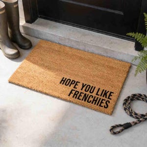 A textured door mat with the phrase "HOPE YOU LIKE FRENCHIES" printed on it, placed on a concrete floor next to a pair of boots and a plant.