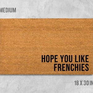 A rectangular, light brown doormat with the text "HOPE YOU LIKE FRENCHIES" printed on it.