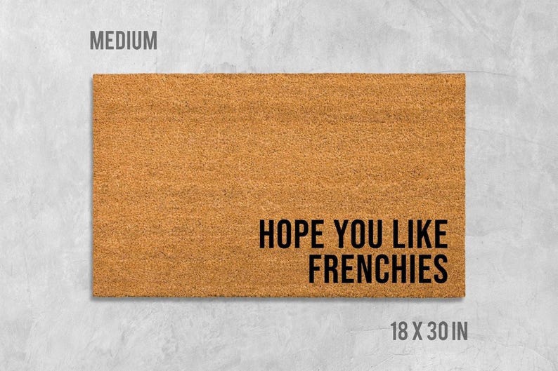 A rectangular, light brown doormat with the text "HOPE YOU LIKE FRENCHIES" printed on it.