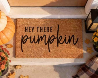 Puede incluir: Un felpudo rectangular de fibra de coco color canela con el texto impreso en negro "HEY THERE pumpkin" en una combinación de fuentes de bloque y cursiva. La alfombrilla está colocada sobre un escalón de hormigón frente a una puerta de madera.