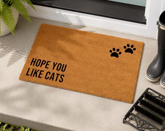 May include: A rectangular tan coir doormat featuring the black text HOPE YOU LIKE CATS on the left side and two black cat paw prints on the right side. The mat has a coarse, fibrous texture and is positioned on a flat concrete surface.