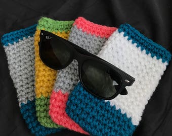 May include: Four crocheted sunglass cases in different color combinations. The cases are made with a striped pattern in gray, white, teal, pink, yellow, and green. A pair of black sunglasses is resting on top of the cases.
