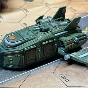 May include: A detailed, green miniature spaceship model with orange and silver accents. The model features wings, a central turret, and intricate paneling. It is set on a tan and brown hexagonal grid.