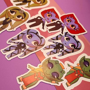 May include: A set of colorful cartoon character stickers featuring a variety of characters, including a green alien, a blue alien, and a green and yellow alien. The stickers are on a pink background.