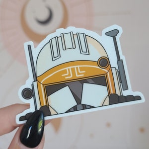 Könnte beinhalten: Ein Aufkleber mit einer Cartoon-Illustration eines weißen und orangefarbenen Helms mit schwarzen und grauen Details. Der Helm hat ein stilisiertes Design mit geometrischen Formen und Linien. Der Aufkleber wird von einer Hand mit einem schwarzen Nagel gehalten.
