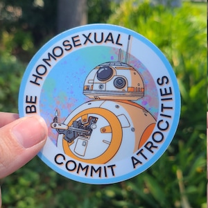 May include: A circular sticker with a cartoon droid design in orange and white, set against a light blue border. The text "BE HOMOSEXUAL COMMIT ATROCITIES" is arranged around the droid. The background is a blurred green.