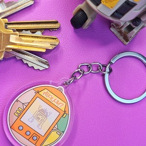 May include: A clear keychain with a silver chain and a round, orange and yellow plastic charm. The charm features a pixelated image of a robot with the text "Tamagotchi" on the screen.