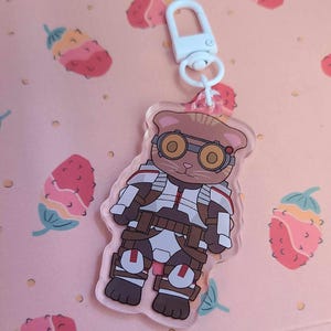 Pode incluir: Um chaveiro de acrílico transparente com um gato de desenho animado usando óculos de proteção e um traje espacial branco e marrom. O chaveiro tem um fecho de metal branco.