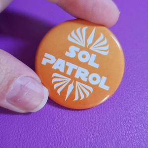May include: Orange button with white text that reads "SOL PATROL" and a white stylized sunburst design.