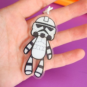 Puede incluir: Un llavero de plástico blanco y negro con un Stormtrooper de dibujos animados con detalles cosidos. El llavero tiene la forma de un juguete de peluche.