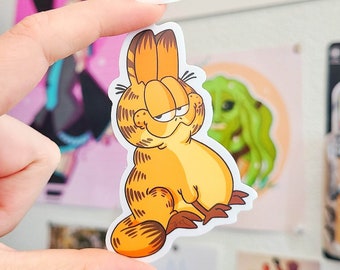 May include: A sticker featuring a cartoon orange tabby cat with a grumpy expression. The cat is sitting with its legs crossed and has a white background.
