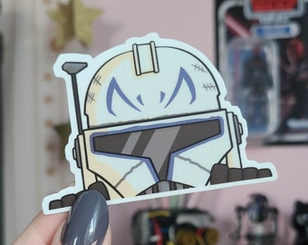 May include: A white and blue cartoon sticker of a Star Wars clone trooper helmet with a blue stripe and stitches on the side. The helmet is looking to the right.
