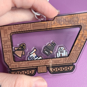 May include: A clear acrylic keychain featuring a brown vehicle design with a series of Star Wars characters inside. The keychain has a white loop at the top for attachment. The background is a solid purple color.
