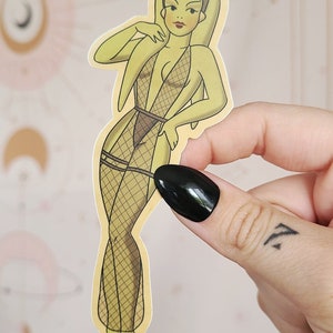Könnte beinhalten: Ein gelber Aufkleber mit einer Cartoon-Illustration einer weiblichen Figur mit grüner Haut, die einen schwarzen und goldenen Netzbody und eine goldene Krone trägt. Die Figur ist mit einer Hand auf der Hüfte und der anderen Hand am Kinn aufgestellt.