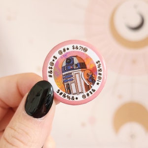 May include: A pink button with a colorful, abstract background featuring a cartoon illustration of R2-D2. The button has a white border with the text "#&$ * $&?!@ $#&%&* @#$& *!@#&%$ ".