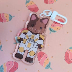 May include: A brown and white cat wearing a white and gold space suit with a white key chain attached. The cat is standing on a pink background with a floral pattern.