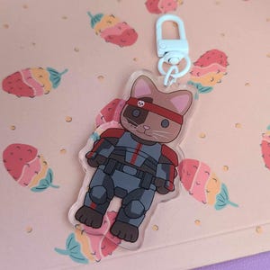 Pode incluir: Um chaveiro de acrílico transparente com um gato de desenho animado usando uma bandana vermelha e um traje espacial cinza e preto. O gato tem pelo marrom e uma mancha branca no rosto.