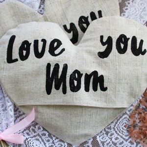 May include: Two heart-shaped linen pouches with black lettering. The pouches say "Love you Mom" and "Love you". The pouches are on a white lace surface with a small bouquet of flowers and pink ribbon.