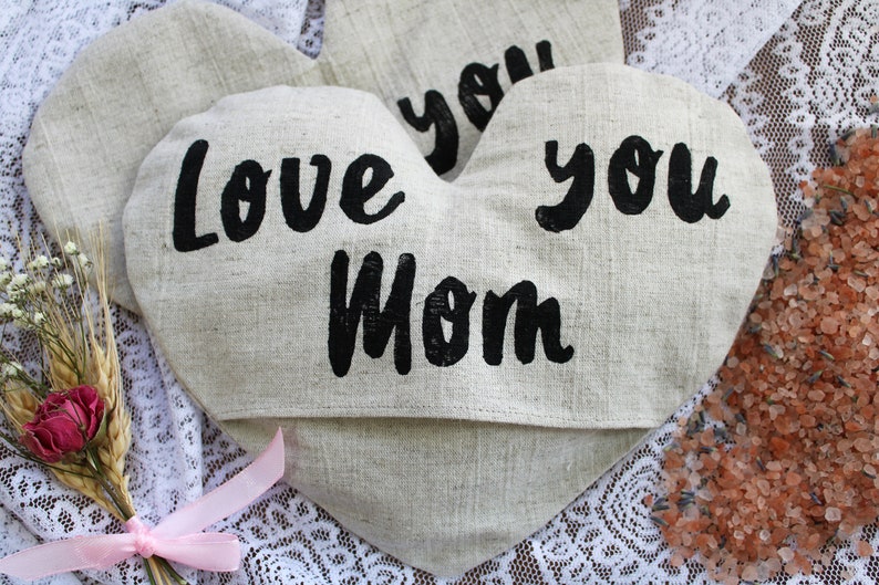 May include: Two heart-shaped linen pouches with black lettering. The pouches say "Love you Mom" and "Love you". The pouches are on a white lace surface with a small bouquet of flowers and pink ribbon.
